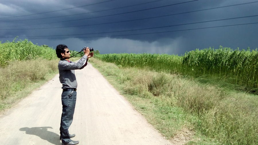 Side view of man standing on road while photographing against cloudy sky using camera
