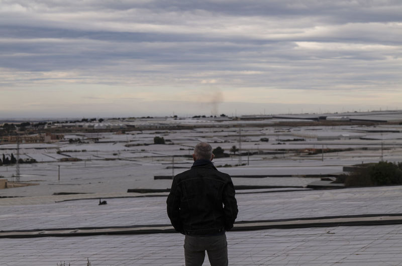 Adult man standing in front of plastic greenhouses against cloudy sky in almeria, spain