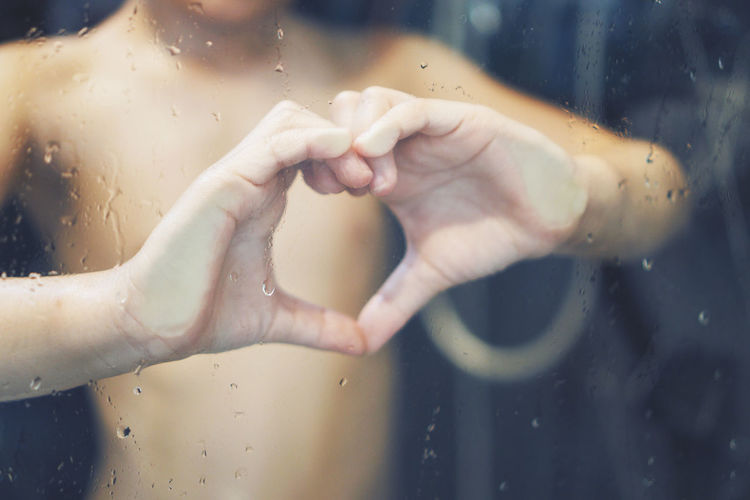 Midsection of shirtless boy making heart shape seen through wet bathroom glass