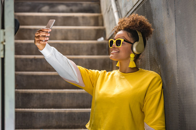 Smiling young woman taking selfie while listening music on mobile phone against wall