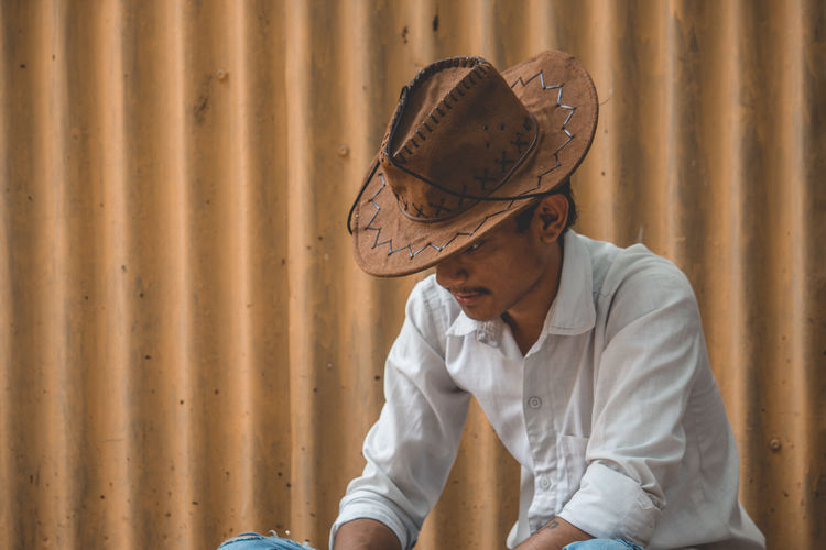 Teenage boy wearing hat sitting against corrugated iron