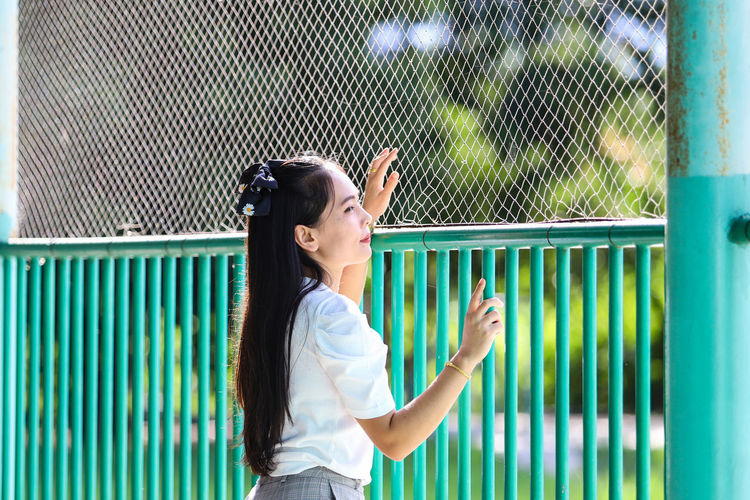Side view of young woman standing against fence
