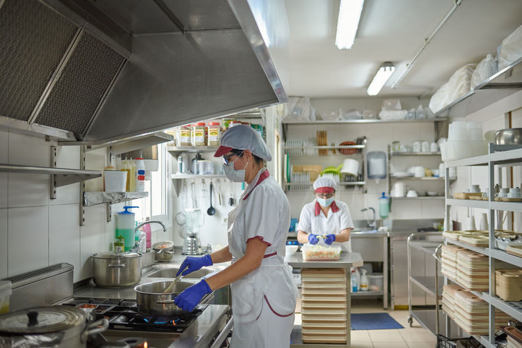 Professional female cook in white uniform and protective mask and gloves preparing food on stove while working with colleague in hospital kitchen during coronavirus pandemic