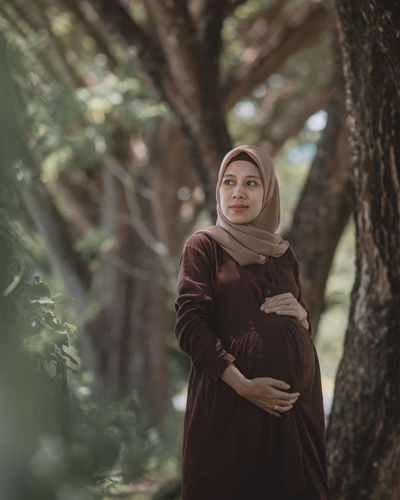 Pregnant woman standing by tree trunk in forest