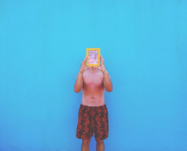 Man holding picture frame in front of face while standing against blue wall