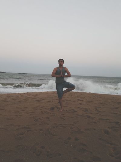 Full length of man doing tree pose yoga while exercising on shore at beach against sky