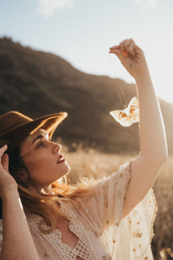 Beautiful woman wearing hat while holding pendant during sunset