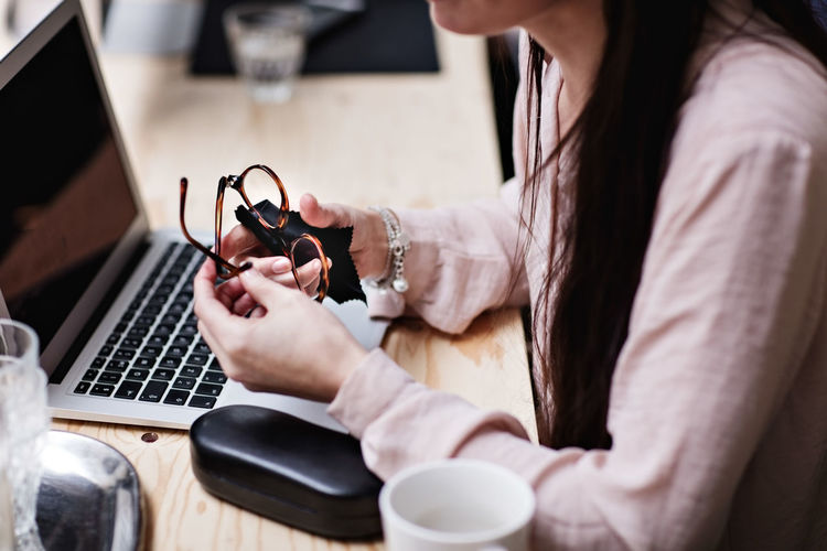 Midsection of businesswoman cleaning eyeglasses by laptop at table in office