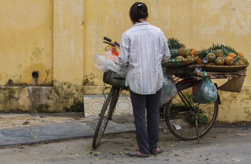 Rear view of vendor standing by pineapples on bicycle against building