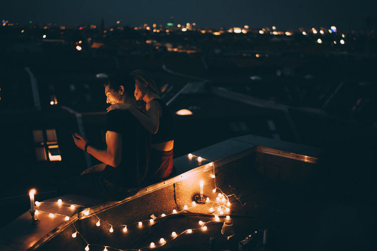 High angle view of couple looking at mobile phone while sitting on illuminated terrace in city at dusk
