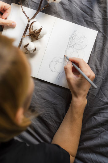 A woman draws a pencil sketch of a cotton twig in a sketchbook.