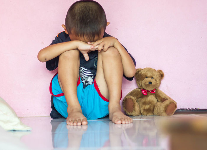 Full length of sad boy sitting by teddy bear on floor at home