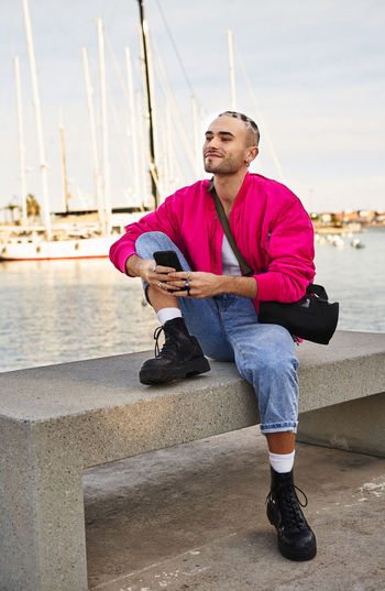 Positive eccentric male with stylish haircut in modern outfit browsing cellphone on marina with moored boats