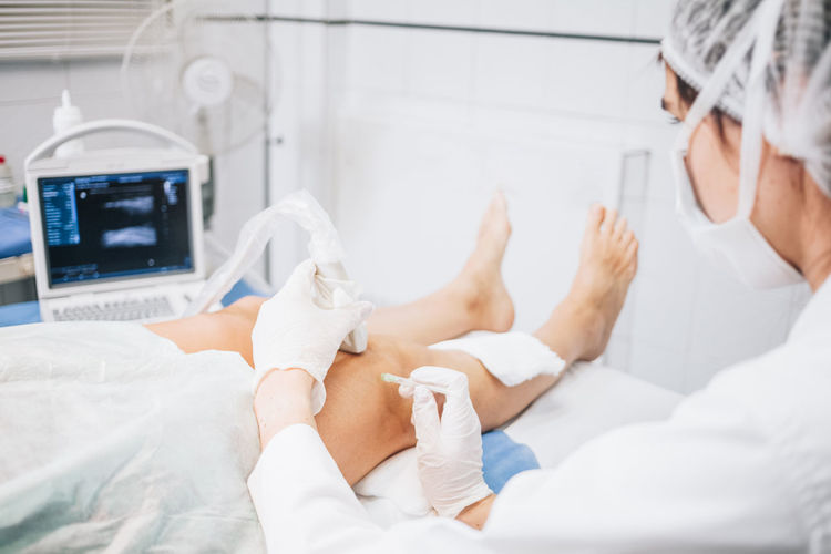 Selective focus on the hands of a doctor wearing a mask, cap, gloves and uniform doing an ultrasound test on the legs of a patient lying on a stretcher in the hospital