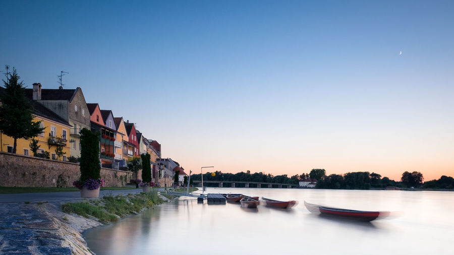 Blurred motion of boats in sea by houses against sky at dawn