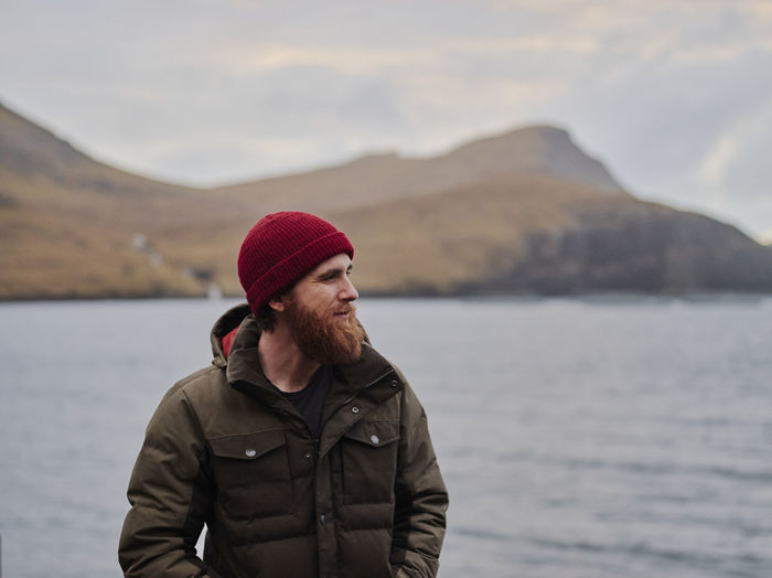 Bearded man in red beanie and jacket standing near ocean